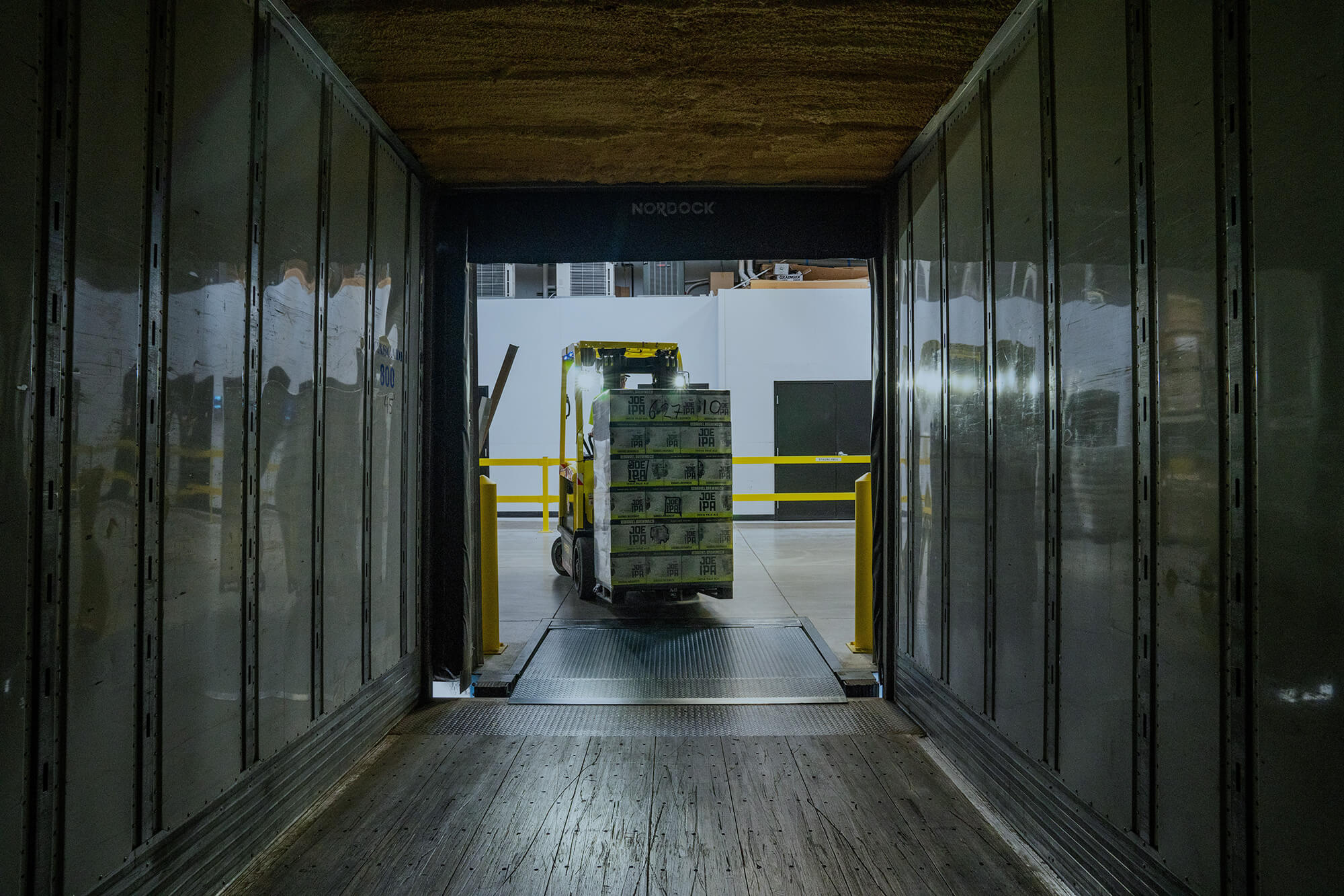 Forklift loading shrink-wrapped pallets into a semi-truck trailer at a warehouse loading dock.